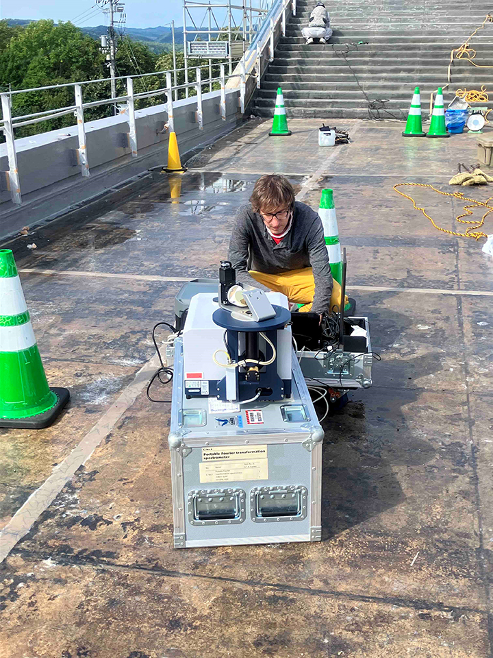 写真1 銀河の森天文台の屋上に設置した可搬型FTIR(EM27/SUN)を用いて太陽光の観測を行っている様子。FTIRを操作しているのは、Matthias Max Frey特別研究員(10月末まで在籍)。屋上の床は、屋根防水補修工事を行っているため、アスファルト防水が剥がされています。
