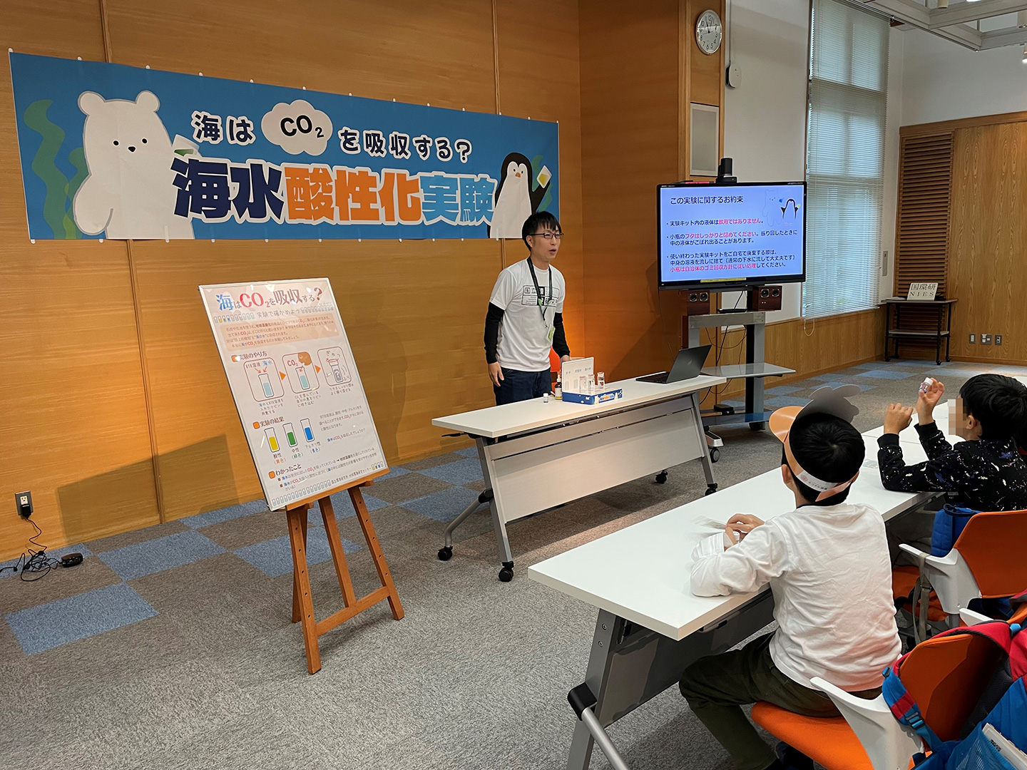 「海はCO2を吸収する？海水酸性化実験」で高尾主任研究員（左）の説明を聞く参加者。参加者全員に海水と指示薬の入った小瓶が配布されます。